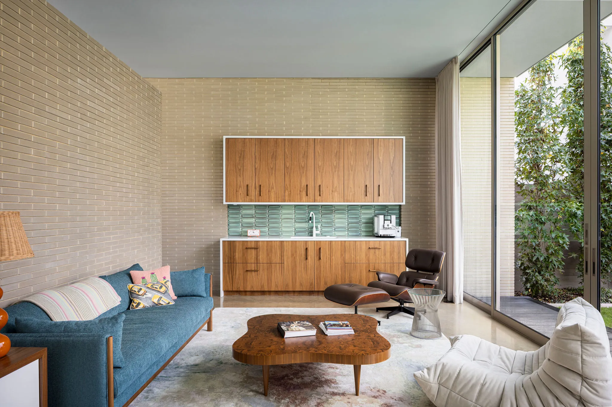 Pool house living room with a teal sofa, walnut kitchenette, lounge chair, and a glass wall to the garden