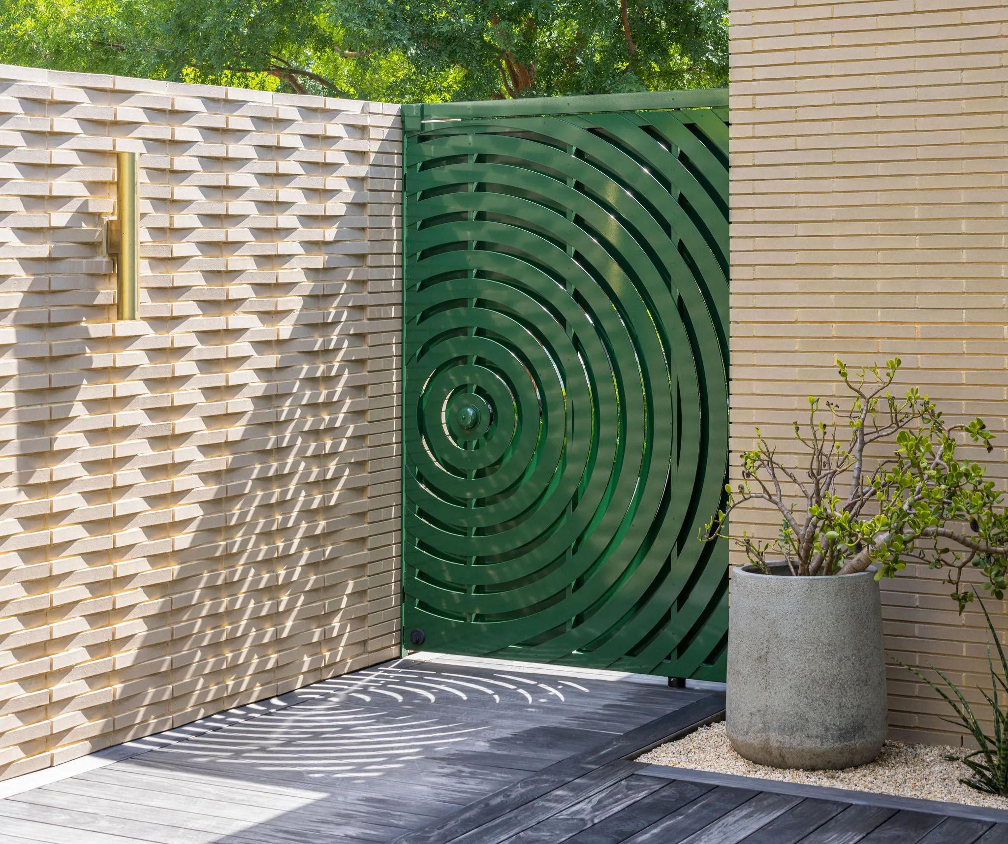 Ripple-pattern green gate casting patterned shadows beside a woven-brick wall and a potted tree