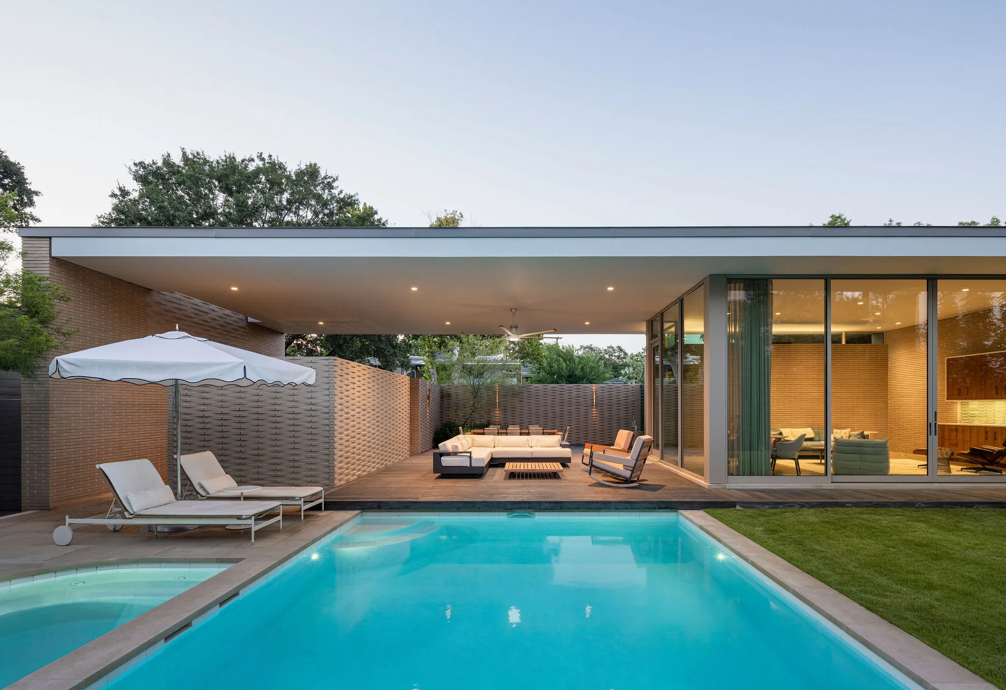 Pool and spa at dusk with loungers, a white umbrella, and a covered terrace under the long flat roof