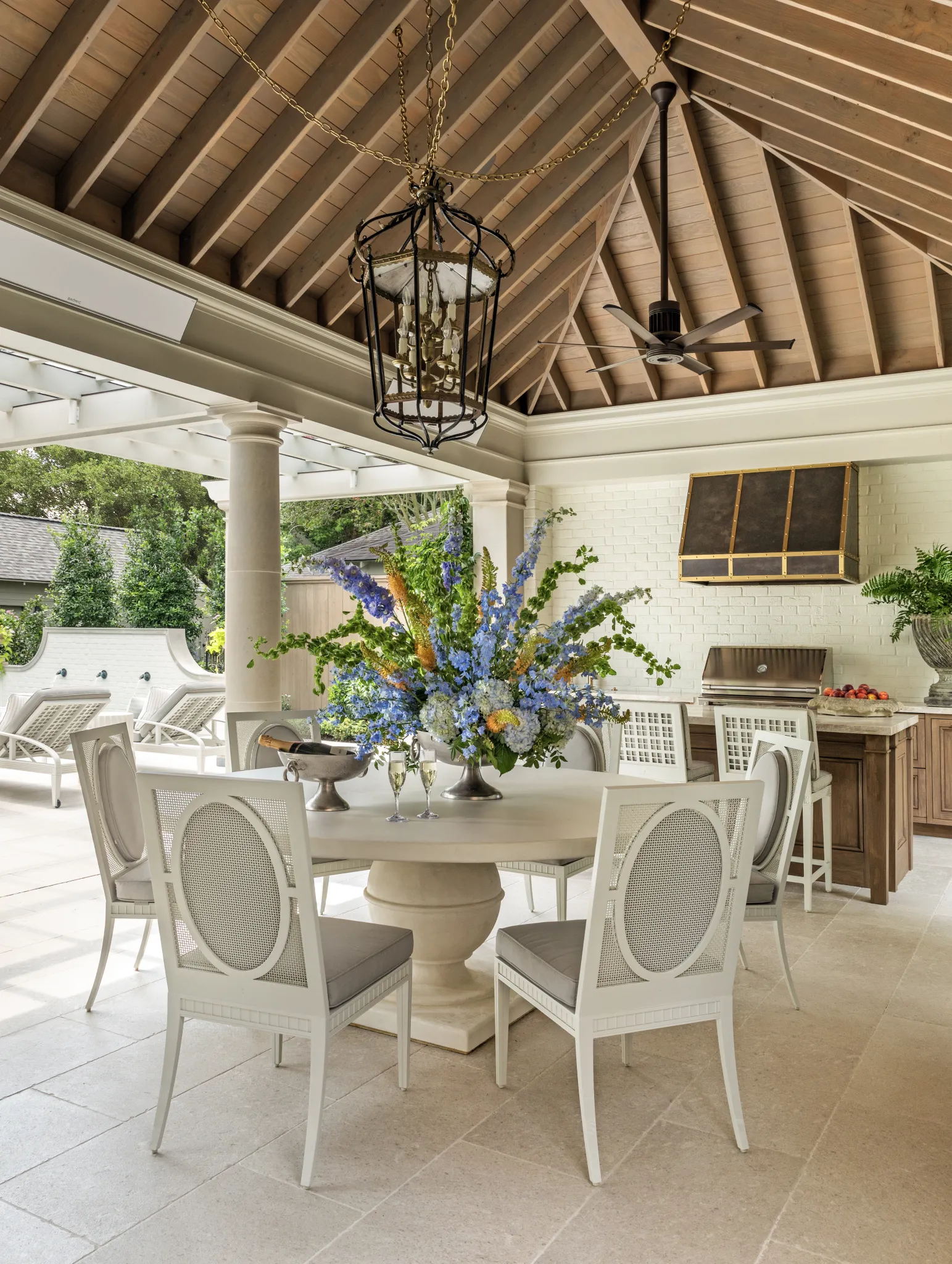 Covered dining room with a round pedestal table, lantern chandelier, and outdoor kitchen beyond