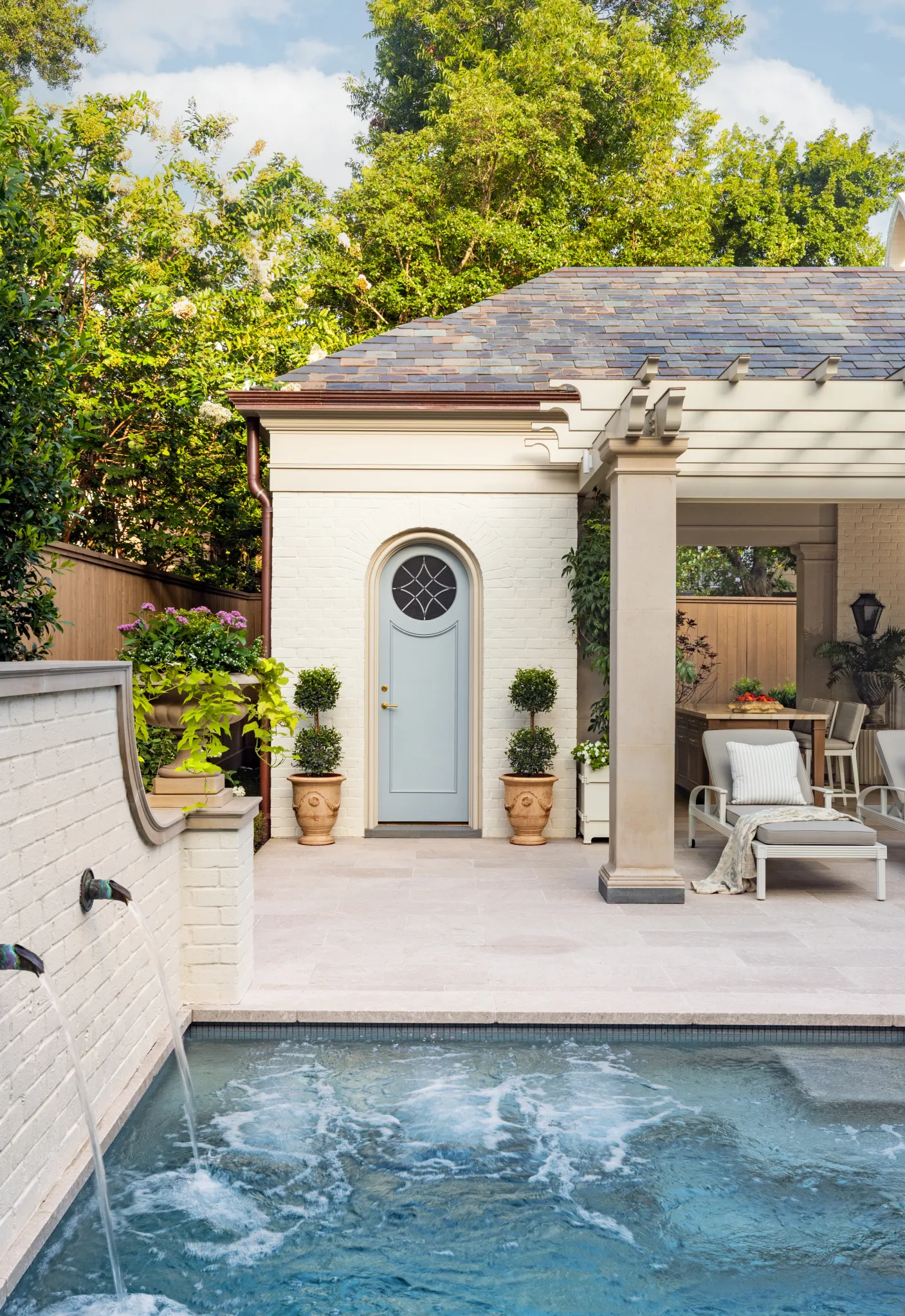 Spa with bronze wall spouts beside the pool house's arched blue door and topiaries